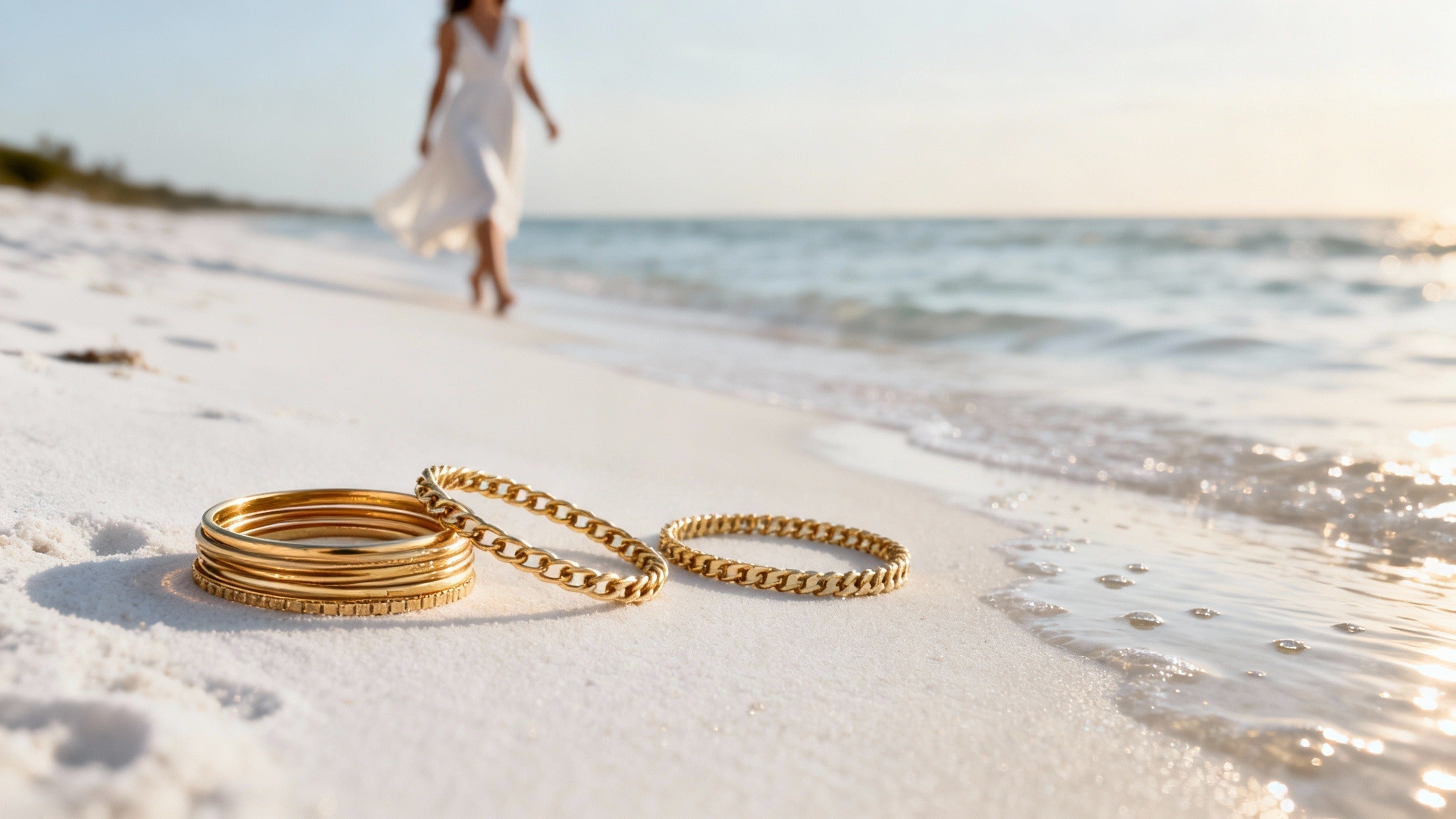 Elevated wristwear for everyday style, featuring neatly arranged gold bangles, chain bracelets, and tennis bracelets on white beach sand with a softly blurred woman walking by the shoreline