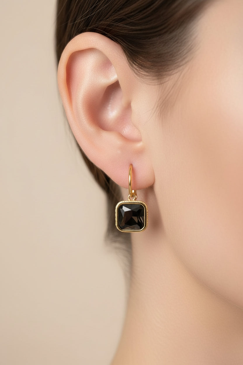 Close-up of square black crystal drop earrings showcasing a faceted black crystal framed in high-polish 18K PVD gold plating.