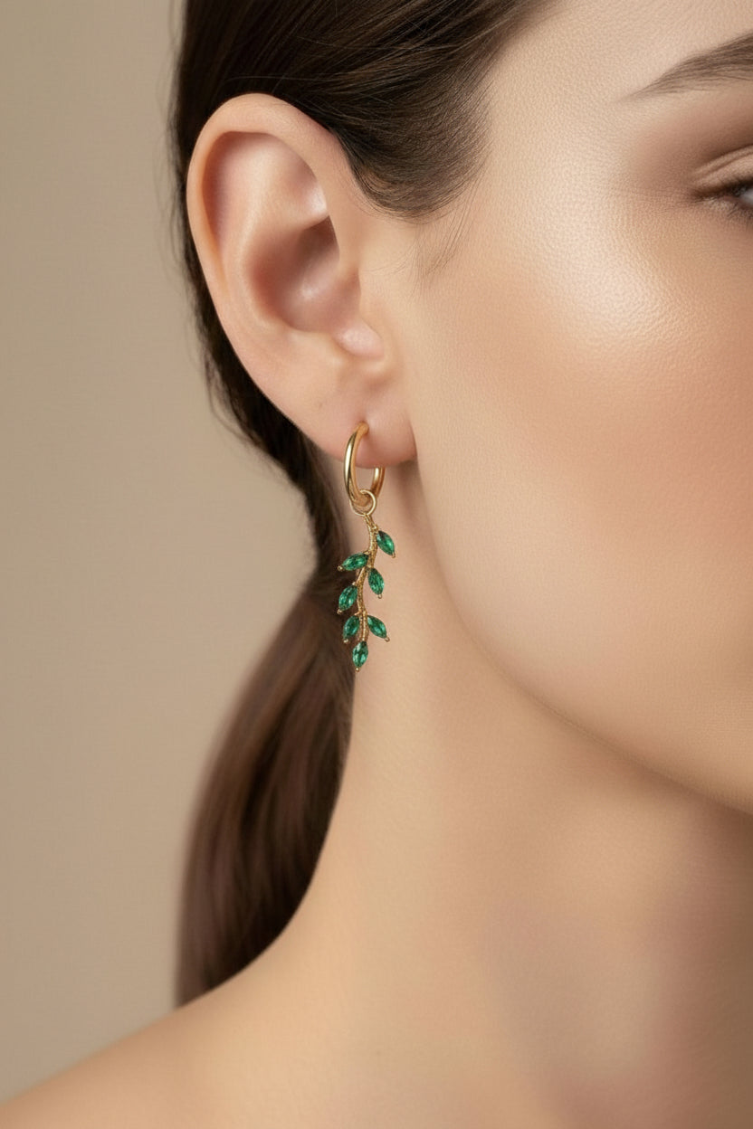 Close-up of green leaf dangle hoop earrings showcasing polished 18K PVD gold plating and marquise-cut green cubic zirconia crystal detailing.