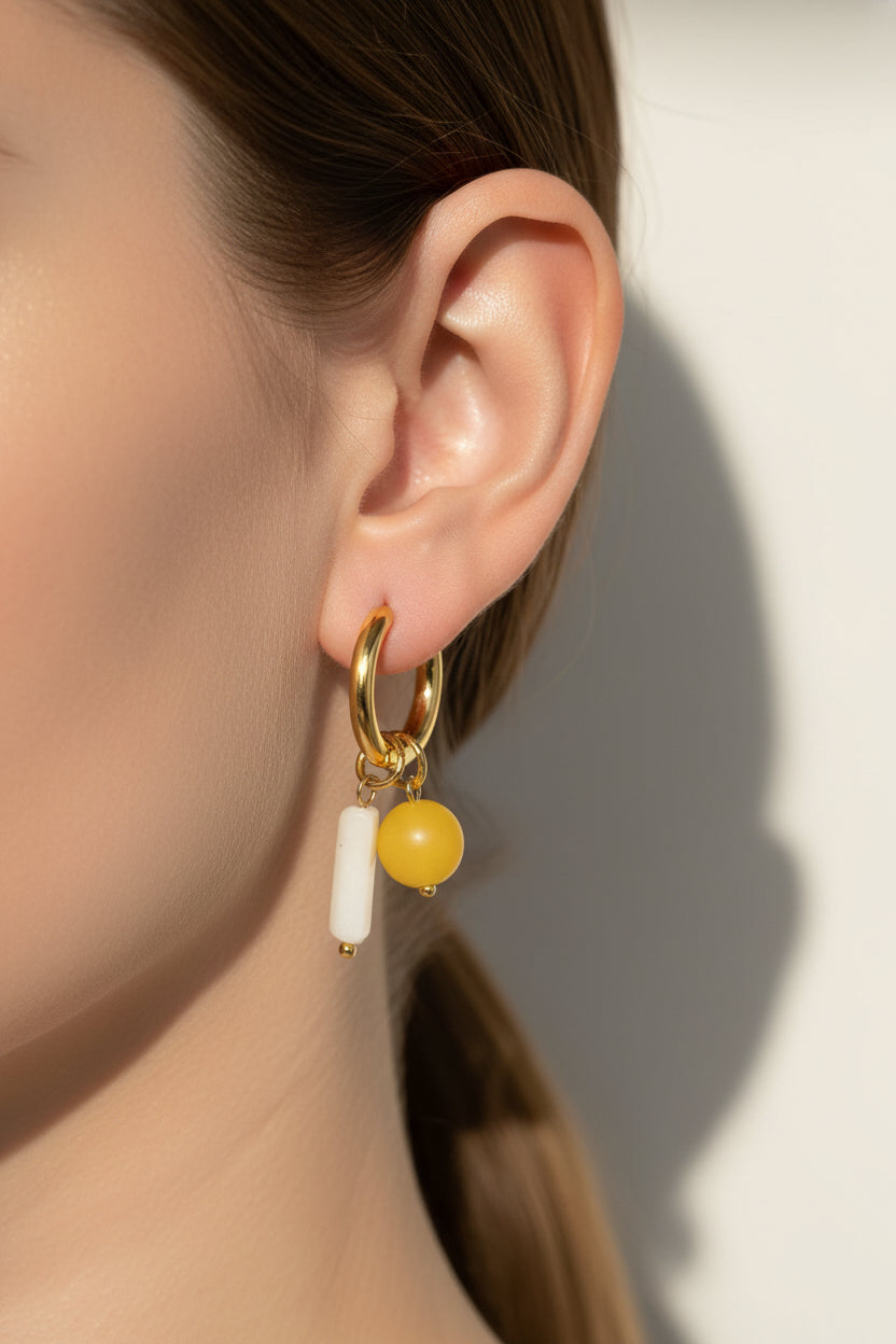 Solara charm hoop drop earrings shown on ear, featuring stainless steel hoops with 18K PVD gold plating and yellow bead and white bar charms.