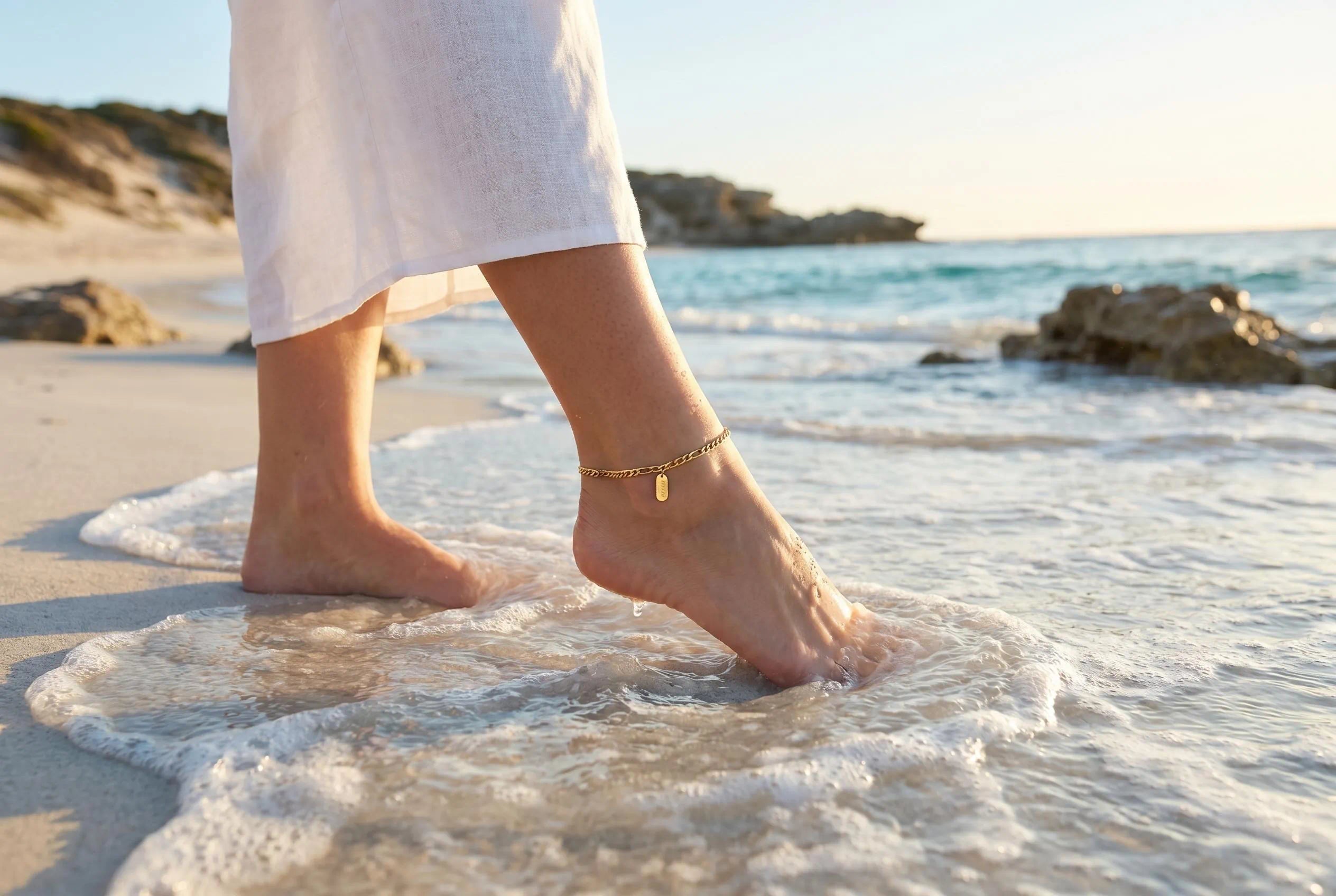Woman walking barefoot on an Australian beach wearing an elegant 18K gold plated waterproof anklet by the shoreline at sunset