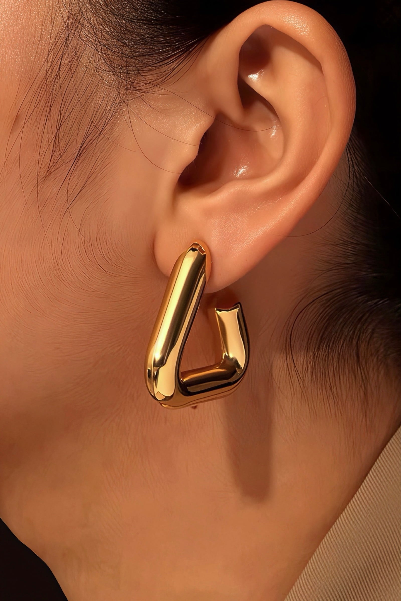 Close-up of a woman wearing geometric triangle hoop earrings made from stainless steel with a high-shine PVD 18K gold plated finish, showcasing the sculptural and modern shape – GLISTIA