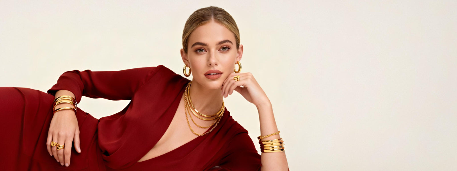 Female model wearing GLISTIA 18K PVD gold plated jewellery including hoop earrings, layered necklaces, bangles and rings styled with a deep red dress against a soft cream background.