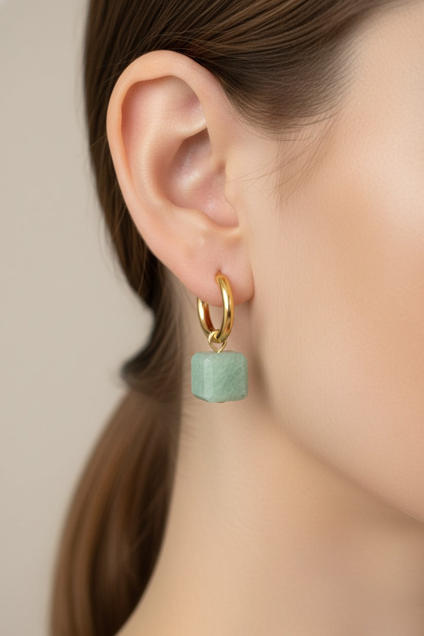 Solara charm hoop drop earrings worn on ear, crafted from stainless steel with 18K PVD gold plating and a green cube charm.
